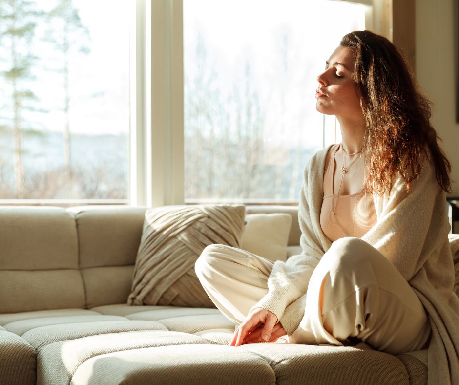 Eine junge Frau sitzt auf einem Sofa, genießt die Sonne, die in ihr Zimmer scheint und entspannt sich.